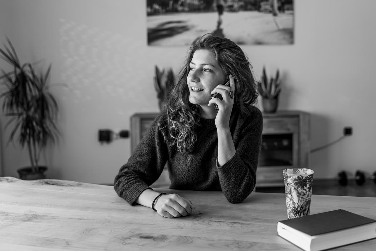 Lächelnde Frau telefoniert an einem Holztisch, daneben ein Buch und ein Glas